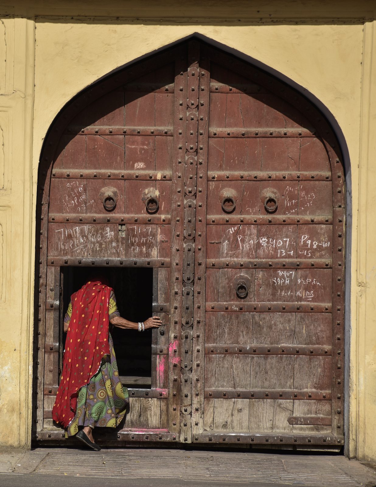 A woman in a red scarf stepping through a studded wooden door, Jaipur